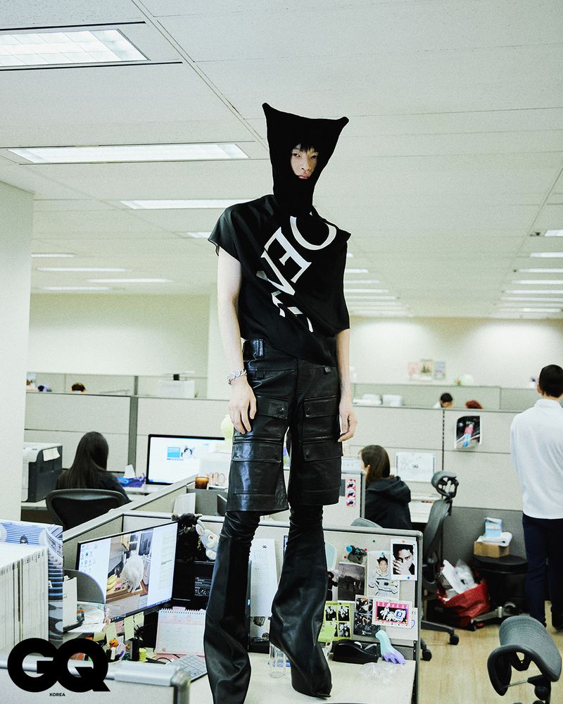 Person with black head covering and T-shirt poses on office desk, wearing black shorts and boots, surrounded by office cubicles and workers.