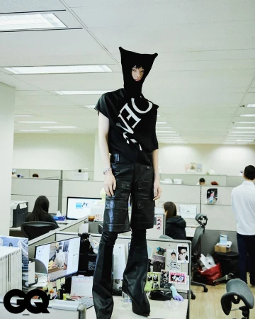 Person with black head covering and T-shirt poses on office desk, wearing black shorts and boots, surrounded by office cubicles and workers.