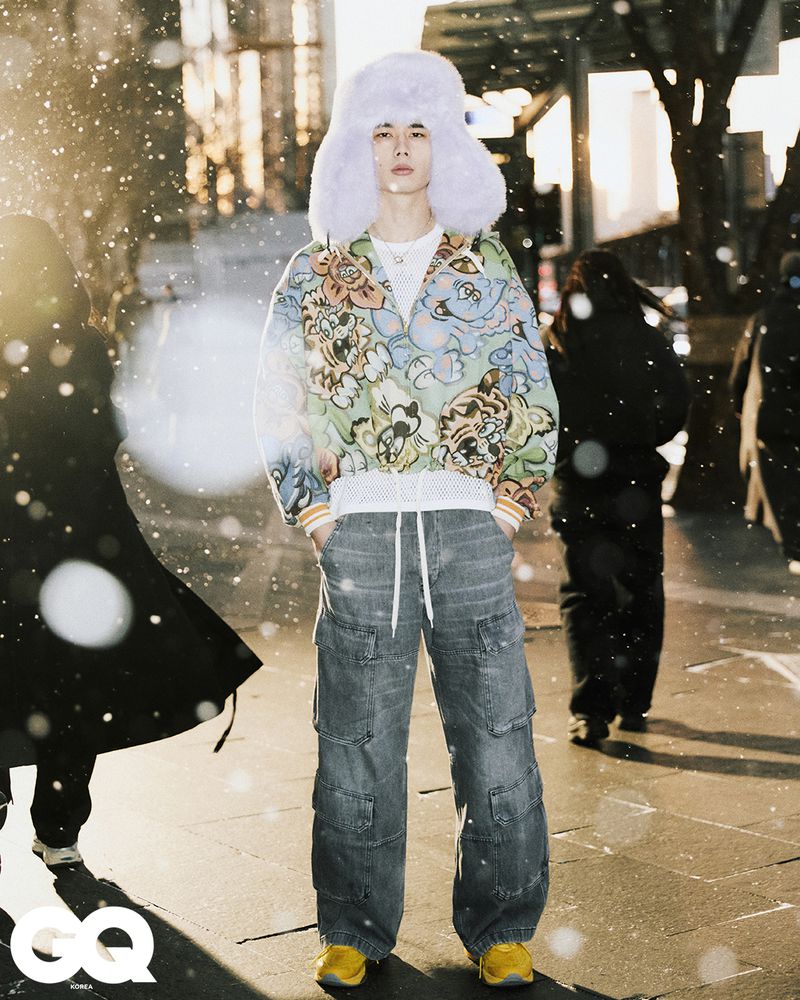 Young adult in patterned jacket, fur hat, and baggy jeans stands on city street with blurred pedestrians and snow effect.