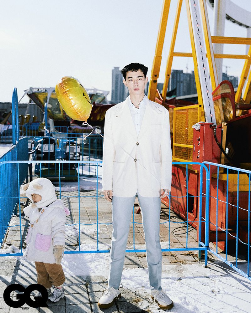 Asian man in suit poses with child holding a balloon in a snowy amusement park, creating a lifestyle editorial fashion shot.