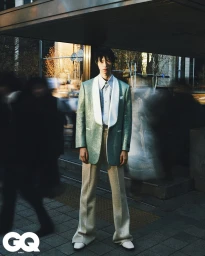 A man in a sparkling green tuxedo jacket and wide trousers standing on a city street with blurred passersby.