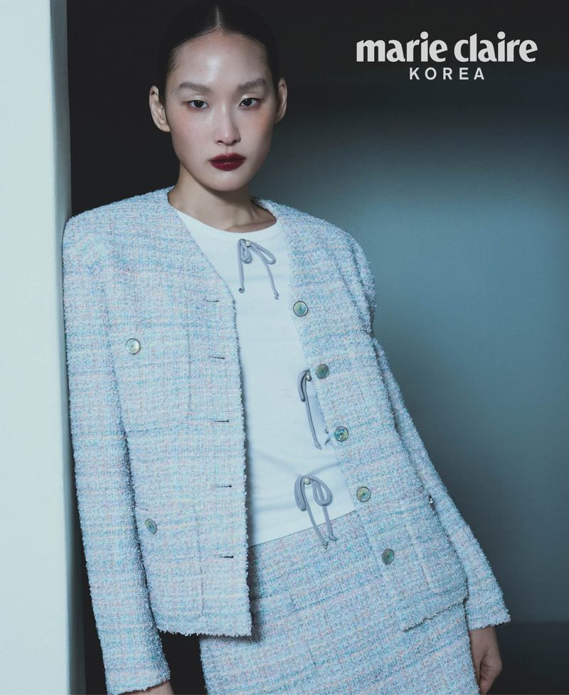 Elegant woman poses in a light blue tweed skirt suit with a white top in a studio. The image features a fashion editorial style.