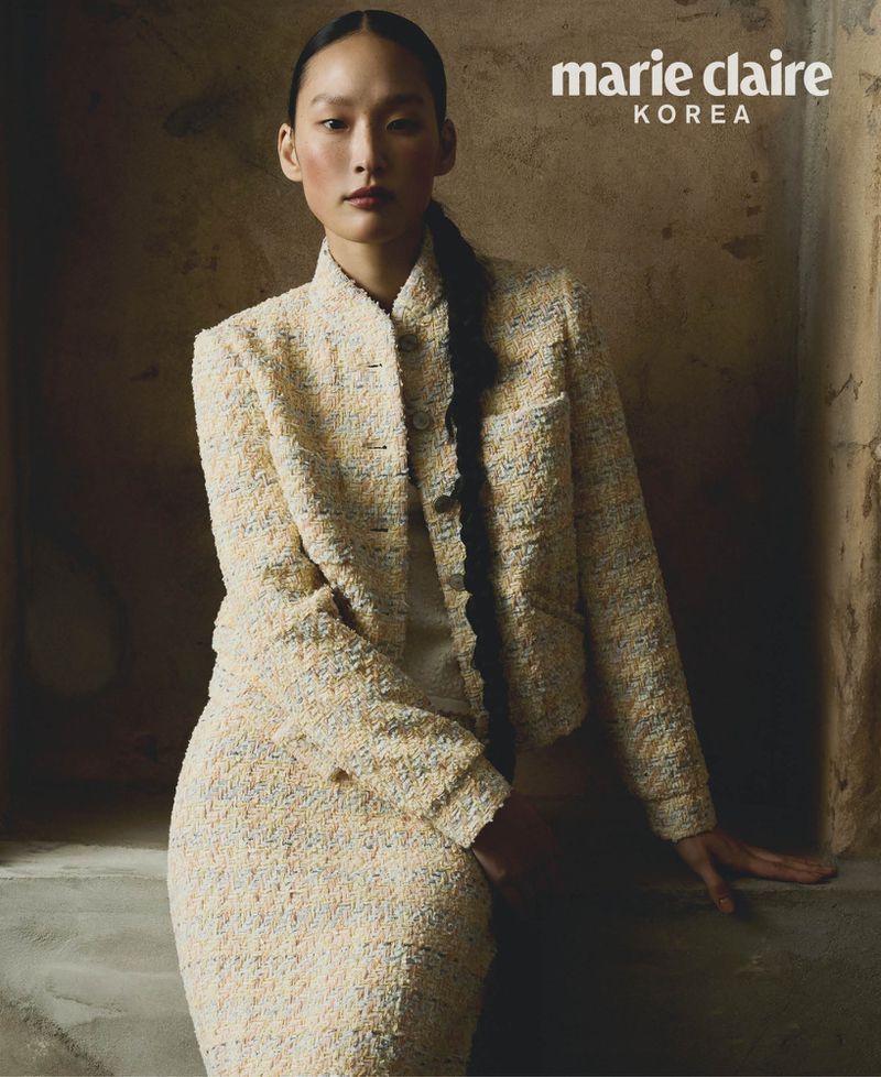 Asian woman poses in a cream-colored tweed skirt suit in a studio with natural lighting, wearing her hair in a long braid.