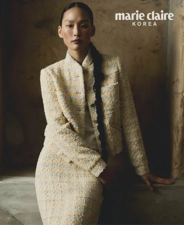 Asian woman poses in a cream-colored tweed skirt suit in a studio with natural lighting, wearing her hair in a long braid.
