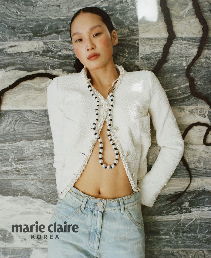 Editorial fashion shot of a slender Asian woman wearing a white jacket and jeans while reclining against a marble tile wall in a studio.