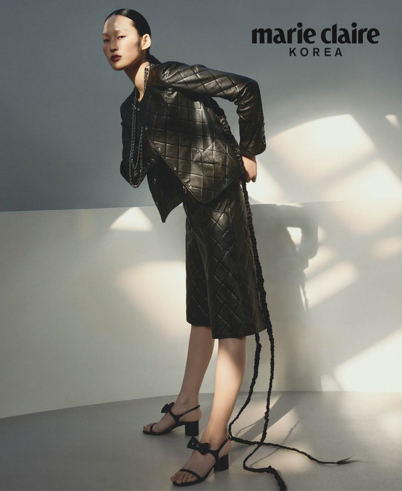 Editorial fashion shot of an Asian woman wearing a black quilted jacket and skirt, accessorized with strappy heels and a chain necklace.