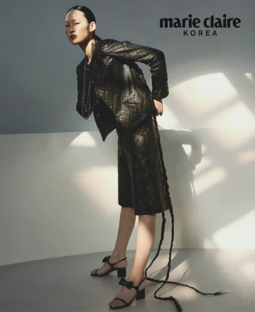 Editorial fashion shot of an Asian woman wearing a black quilted jacket and skirt, accessorized with strappy heels and a chain necklace.