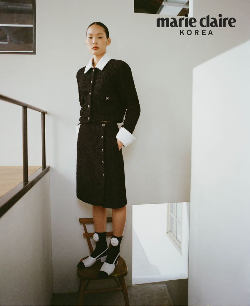 A young Asian female model poses indoors on a vintage wooden chair, wearing a black tweed skirt and jacket outfit in a fashion editorial.