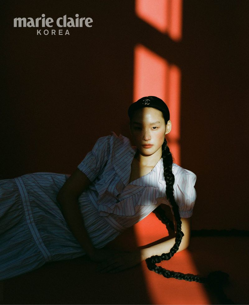 Model with long braided hair wearing a striped dress reclining against a red wall with sharp sunlight shadows for Marie Claire Korea.
