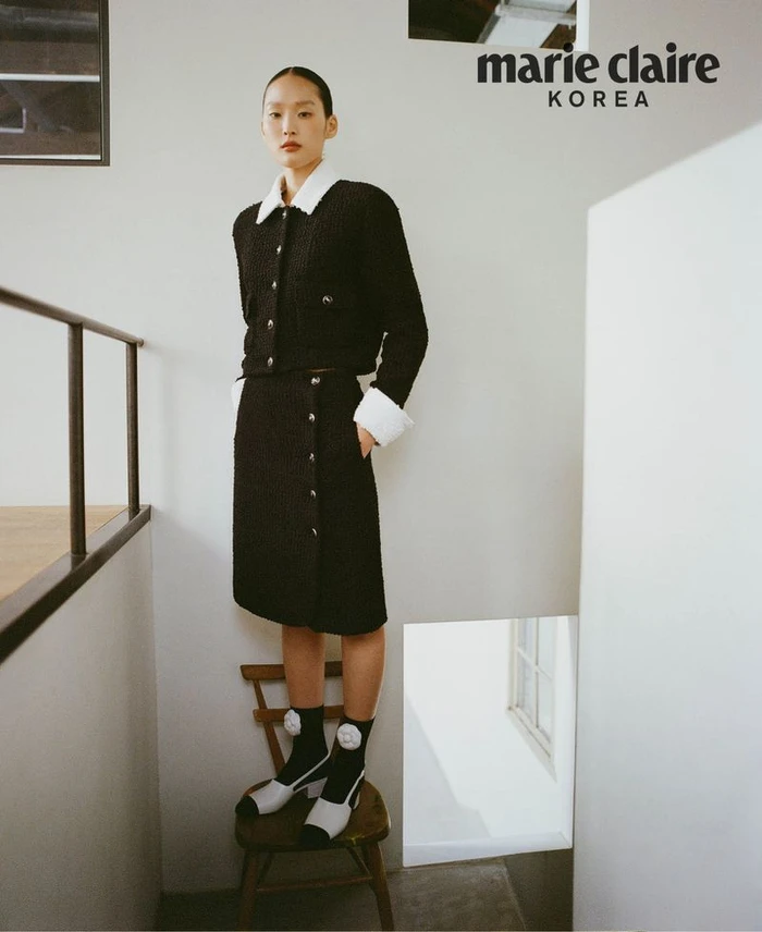 Model standing on a wooden chair wearing a black tweed jacket and skirt set with white trim, for Marie Claire Korea