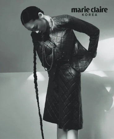 Model wearing a black quilted leather jacket and skirt with a long braided hair extension against a bright background.