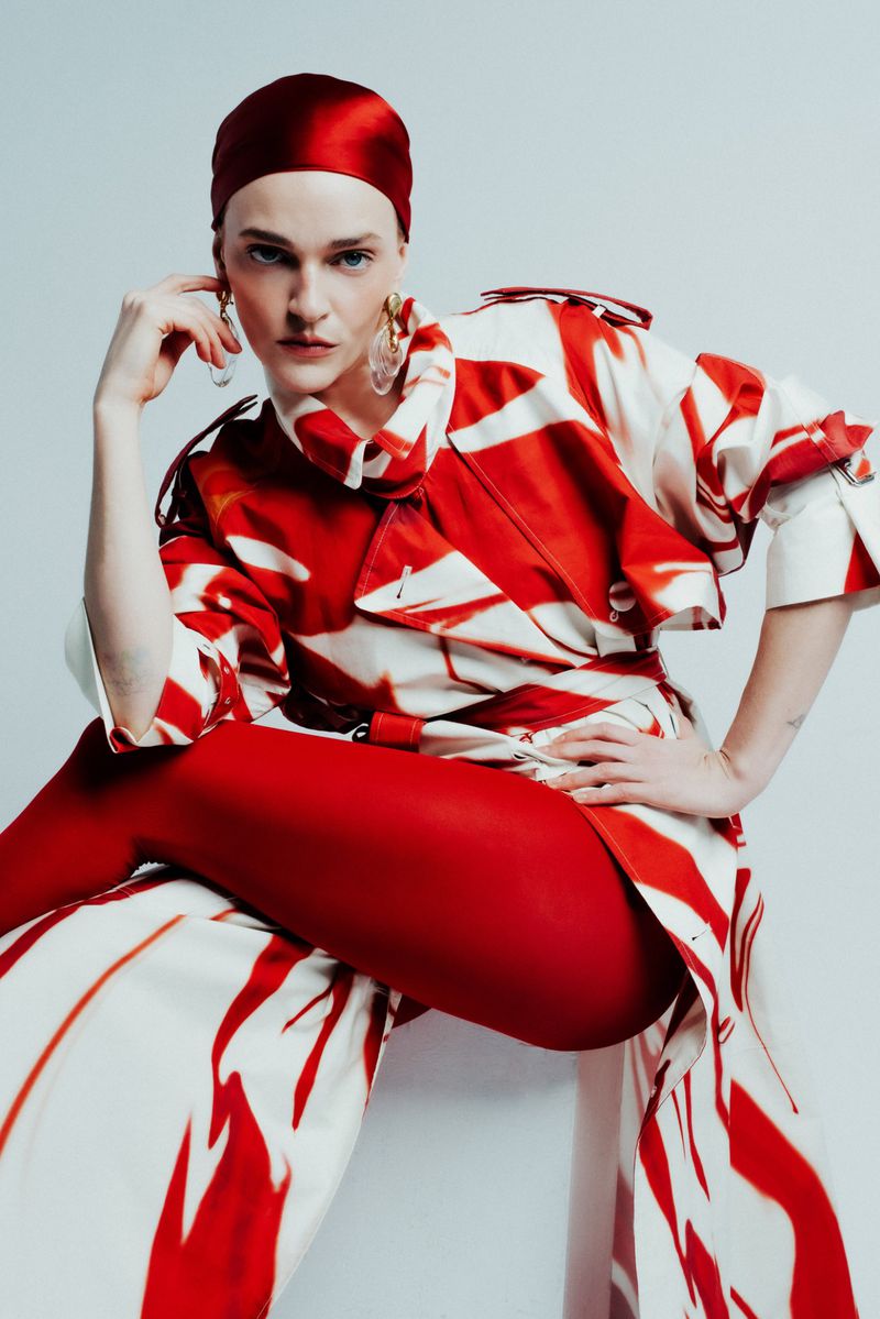 Adult woman poses in studio wearing a red headscarf, oversized patterned coat, and red tights against a plain, light-colored background.