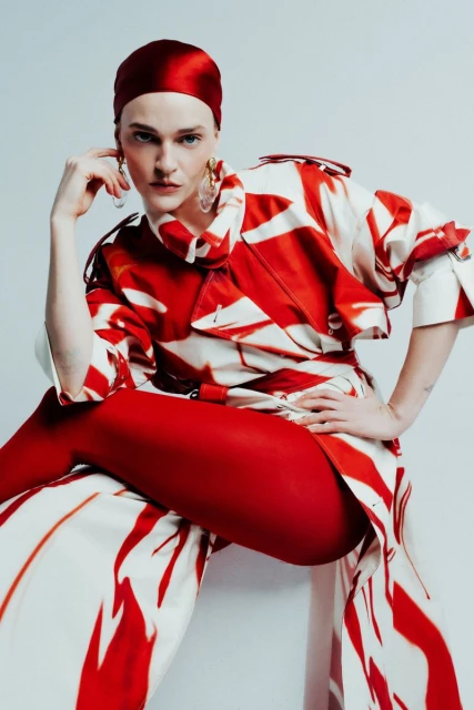 Adult woman poses in studio wearing a red headscarf, oversized patterned coat, and red tights against a plain, light-colored background.