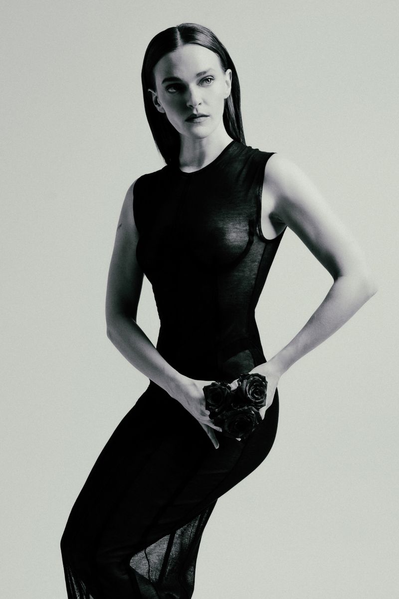 A fair-skinned adult woman in a black dress poses in a photo studio, holding roses, in a fashion editorial portrait, with an elegant and serious expression.
