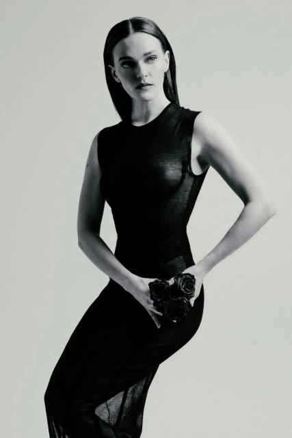 A fair-skinned adult woman in a black dress poses in a photo studio, holding roses, in a fashion editorial portrait, with an elegant and serious expression.