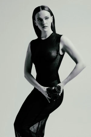 A fair-skinned adult woman in a black dress poses in a photo studio, holding roses, in a fashion editorial portrait, with an elegant and serious expression.