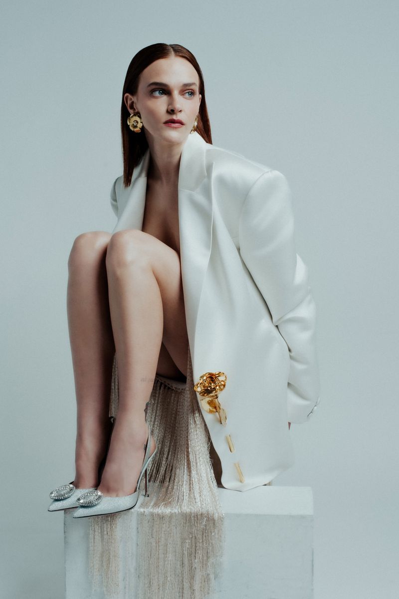 A redhead woman sits on a white cube wearing a white blazer, gold earrings, and silver glittery heels in a fashion style pose.
