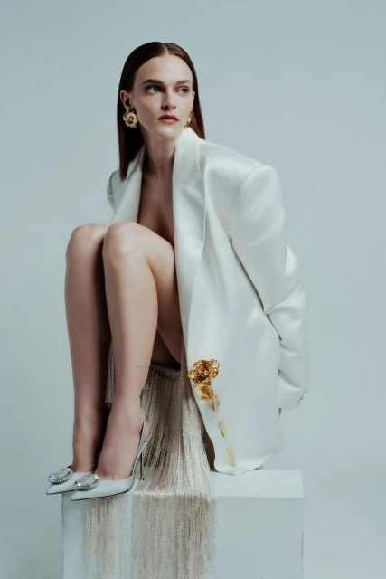 A redhead woman sits on a white cube wearing a white blazer, gold earrings, and silver glittery heels in a fashion style pose.