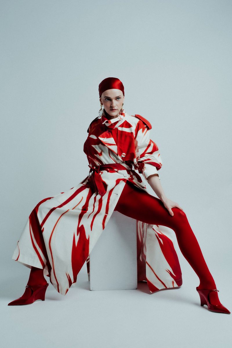 Fashionable woman in patterned coat, red leggings and headdress poses on a white cube against a pale backdrop, showcasing studio style.