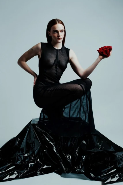 Elegant woman in a sheer black dress poses with red roses in a fashion forward studio shot with a neutral background and dramatic lighting.