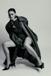 Model Madeline Brewer seated on a chair wearing a dark trench coat and heels against a white studio background