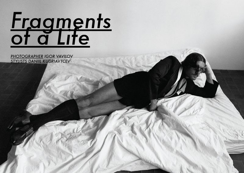 Black and white studio shot of a reclining adult male with dark hair, wearing black jacket and boots, on a bed with white sheets.