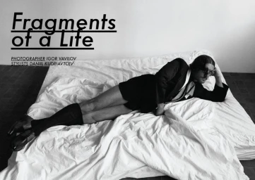 Man lounging on a white unmade bed wearing a dark suit jacket and shorts, photographed in monochrome.