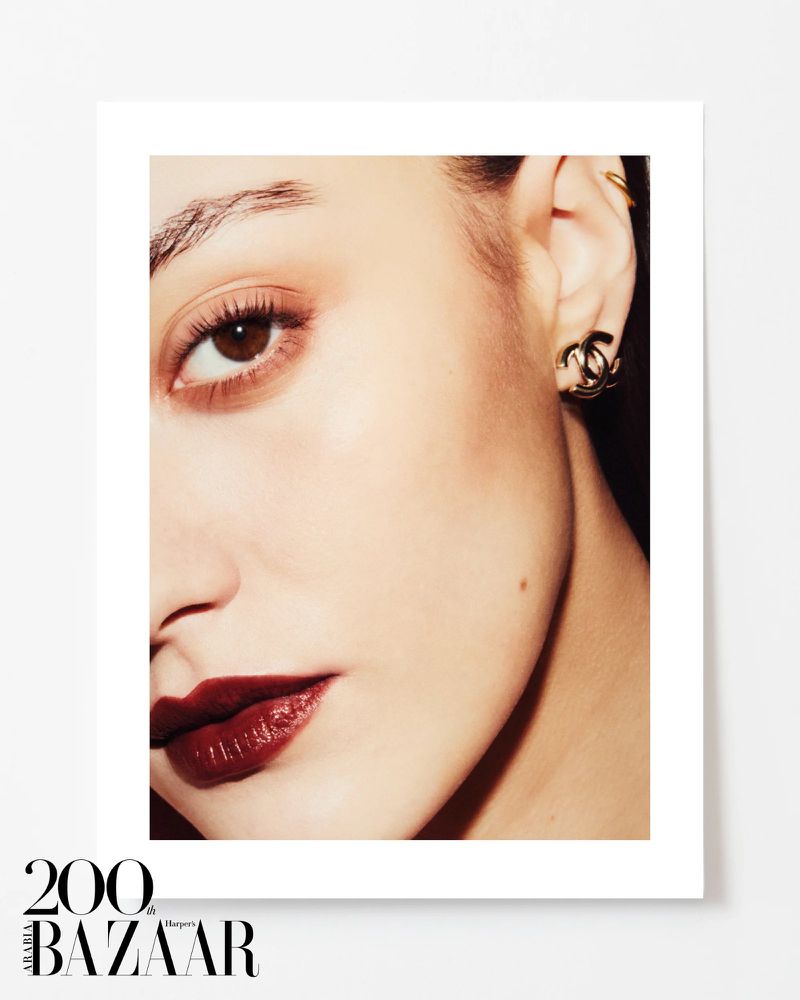 Close-up studio portrait of a young adult woman with dark red lipstick, jewelry, and makeup, against a light neutral background.