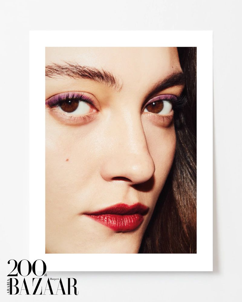 Close-up portrait of a woman with brown eyes, purple eye shadow, dark red lipstick, and dark hair, against a white background.