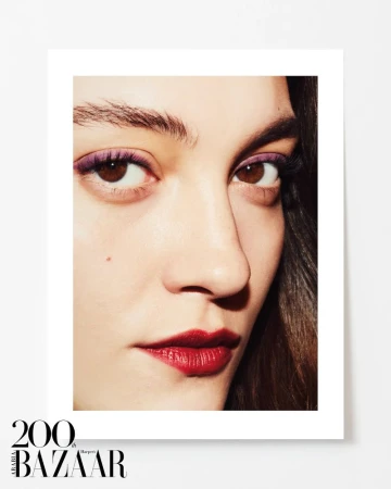 Close-up portrait of a woman with brown eyes, purple eye shadow, dark red lipstick, and dark hair, against a white background.