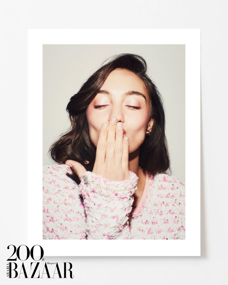 Close-up portrait of a young woman with dark hair, wearing a pink jacket, blowing a kiss in front of a white background.