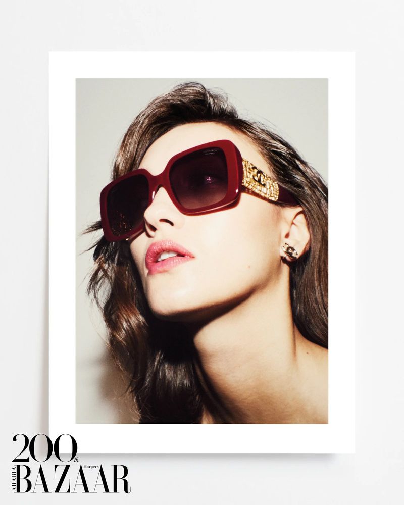 Close-up fashion shot of a woman wearing oversized burgundy sunglasses with gold details and jewelry against a neutral backdrop.
