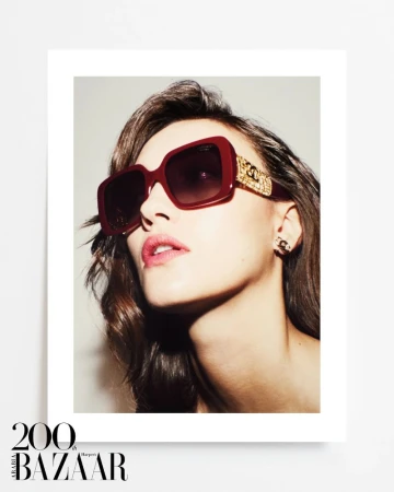 Close-up fashion shot of a woman wearing oversized burgundy sunglasses with gold details and jewelry against a neutral backdrop.