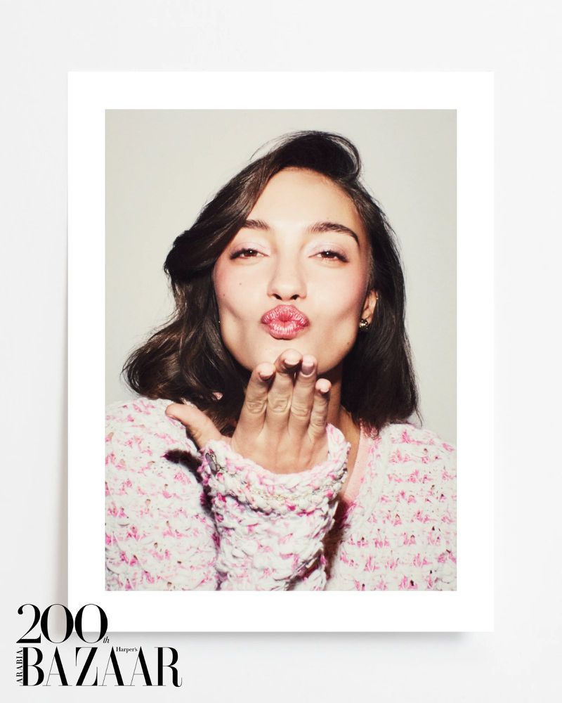 Portrait of a woman with brown hair and pink lipstick, blowing a kiss towards the camera, wearing a pink tweed jacket in a photo studio.