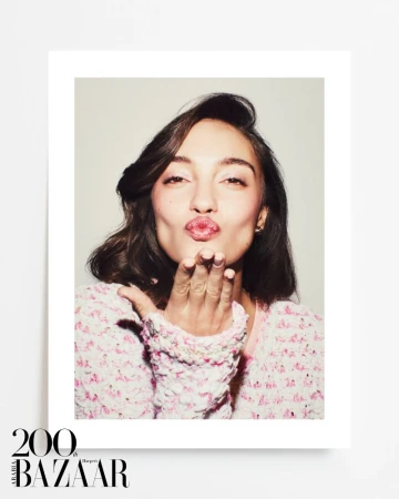 Portrait of a woman with brown hair and pink lipstick, blowing a kiss towards the camera, wearing a pink tweed jacket in a photo studio.