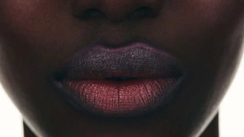 Close-up on an adult female face, highlighting lips adorned with dark red and purple lipstick in a beauty and fashion context.