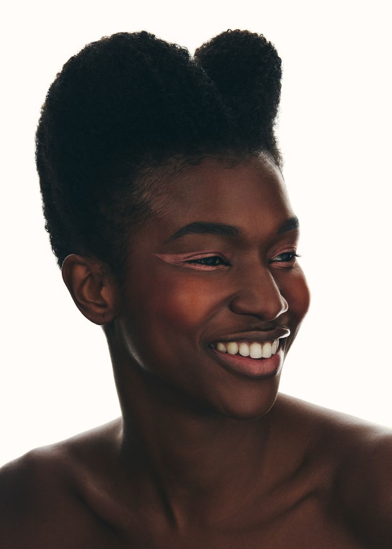 Close-up portrait of a smiling African American woman with stylish dark hair and light pink eyeliner against a white background.