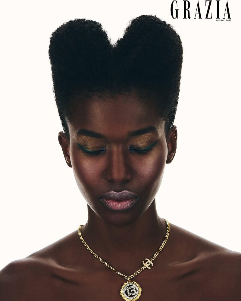 Dark-skinned woman poses in a studio with black heart-shaped afro hairstyle, gold necklace, and bare shoulders against a white backdrop.