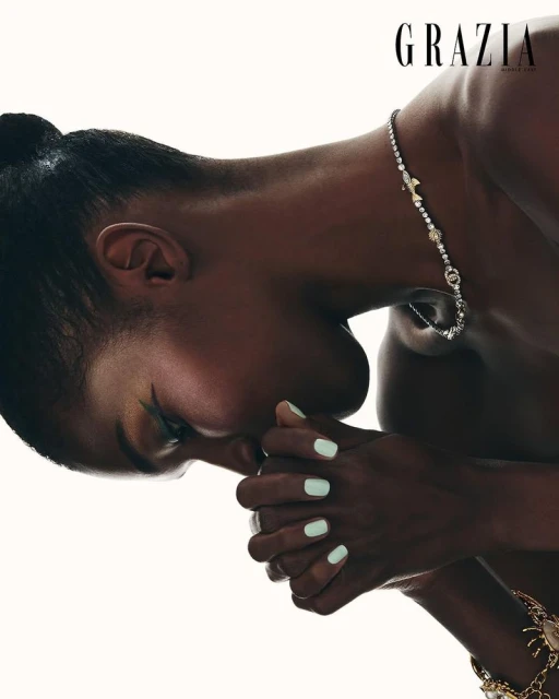Close-up portrait of a woman with dark skin and her hands clasped together, adorned with jewelry, set against a bright white background.