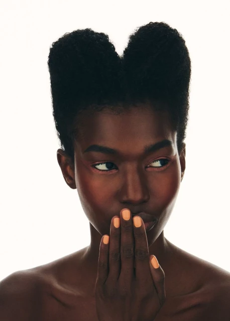 Close-up portrait of a dark-skinned woman with an afro, peach makeup, and coral nail polish posing in front of a white background.