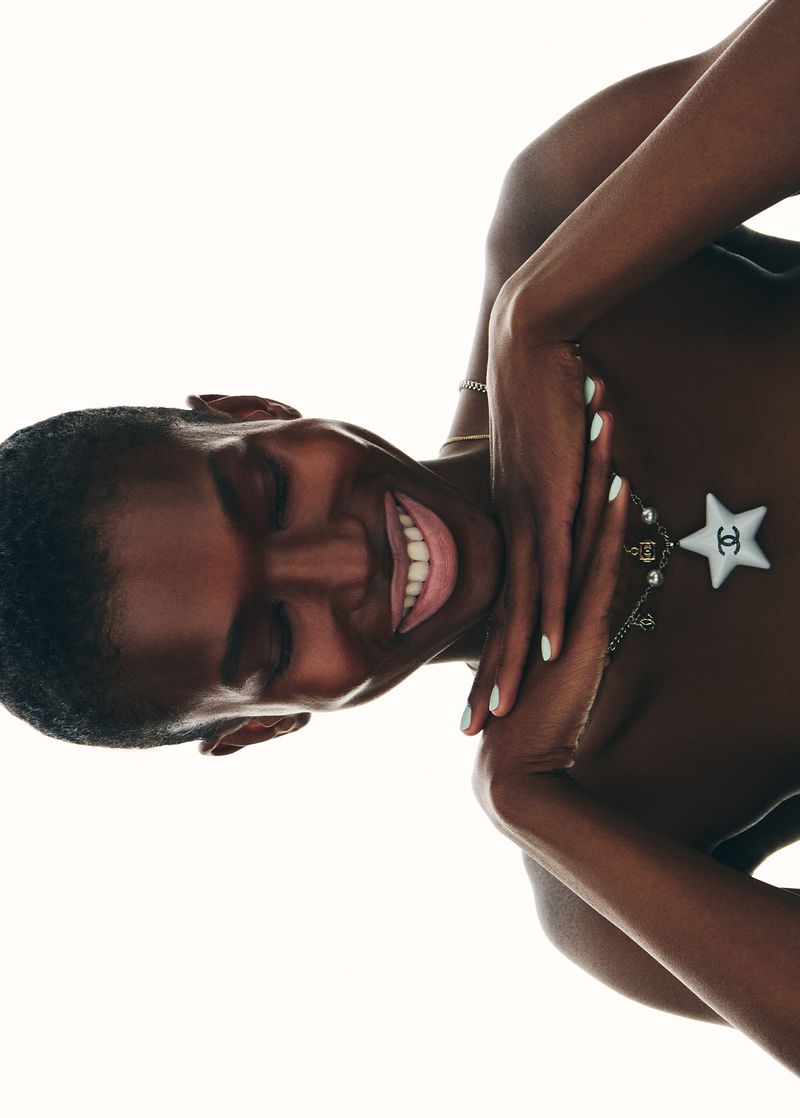 An adult female model with dark skin smiles, lying down against a stark white background, wearing a necklace with a white star pendant.