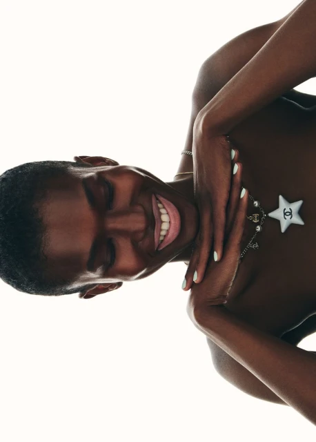 An adult female model with dark skin smiles, lying down against a stark white background, wearing a necklace with a white star pendant.