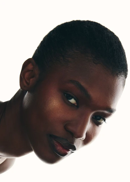 Close-up portrait of a dark-skinned woman with a short haircut, subtle makeup, and glossy lips against a bright, neutral background in soft lighting.