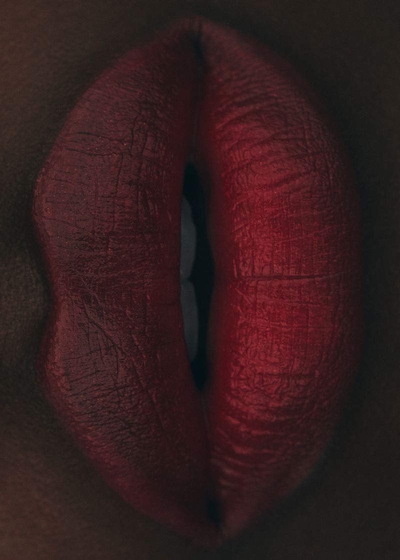Close-up shot of an adult female's red lips, with visible teeth. The woman has dark skin and wears dark red lipstick with a glossy texture.