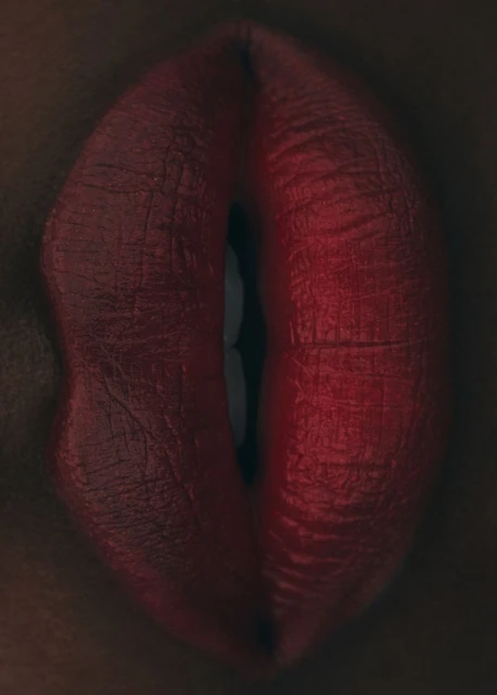 Close-up shot of an adult female's red lips, with visible teeth. The woman has dark skin and wears dark red lipstick with a glossy texture.