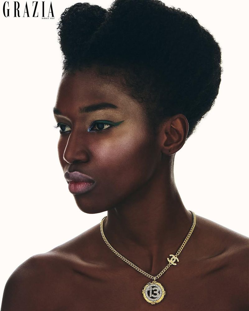 Close-up editorial shot of a Black female model with bold makeup and an afro hairstyle, wearing a gold chain necklace against a white backdrop.