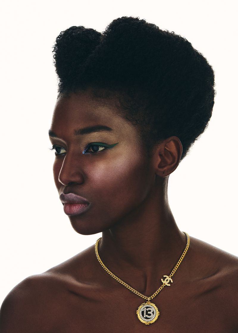 A profile portrait of a Black woman with an afro wearing Chanel jewelry against a white backdrop, showcasing beauty and fashion.