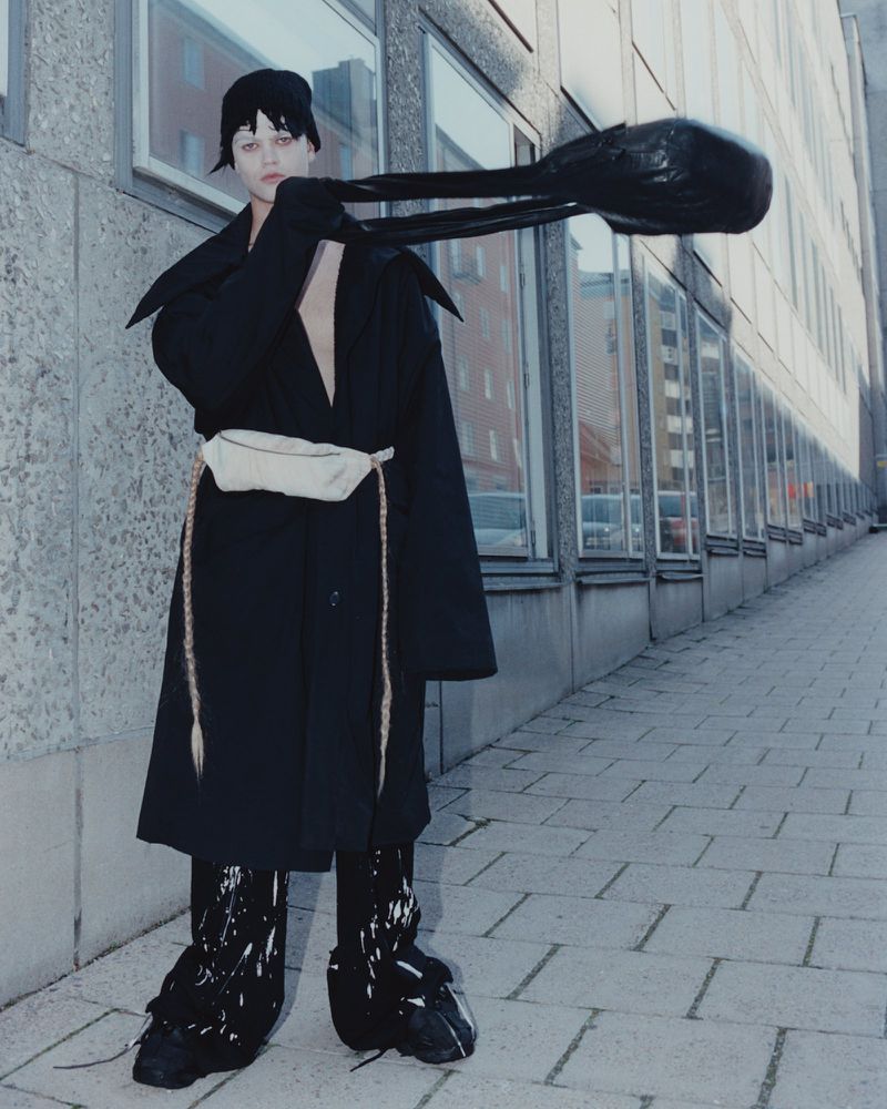 Model in a black coat and hat holding a large black bag on a paved city street