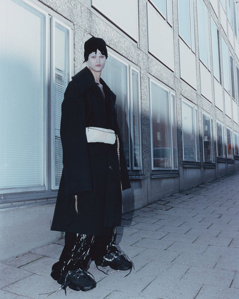 A person stands in front of a building, wearing a black coat, beanie, and unique black shoes, with a beige waist bag and a serious expression.