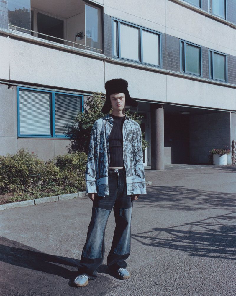 A fair-skinned adult male with a fur hat and patterned shirt stands in front of a building on a sunny day in an urban environment.